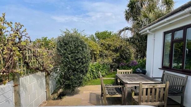 Un jacuzzi dans un jardin, avec une table et des chaises en bois, entouré d’une végétation luxuriante et d’un bâtiment blanc.
