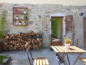 A rustic outdoor seating area with a wooden table and chairs, a stack of firewood, and a shelf with potted plants.