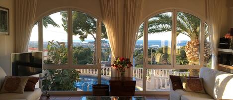 Una sala de estar con una mesa de centro de vidrio, dos sofás, un televisor y ventanas arqueadas amplias con vista a una piscina y paisaje.