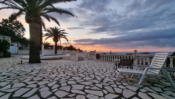 A patio with mosaic flooring, a pool, palm trees, and a view of the sunset.