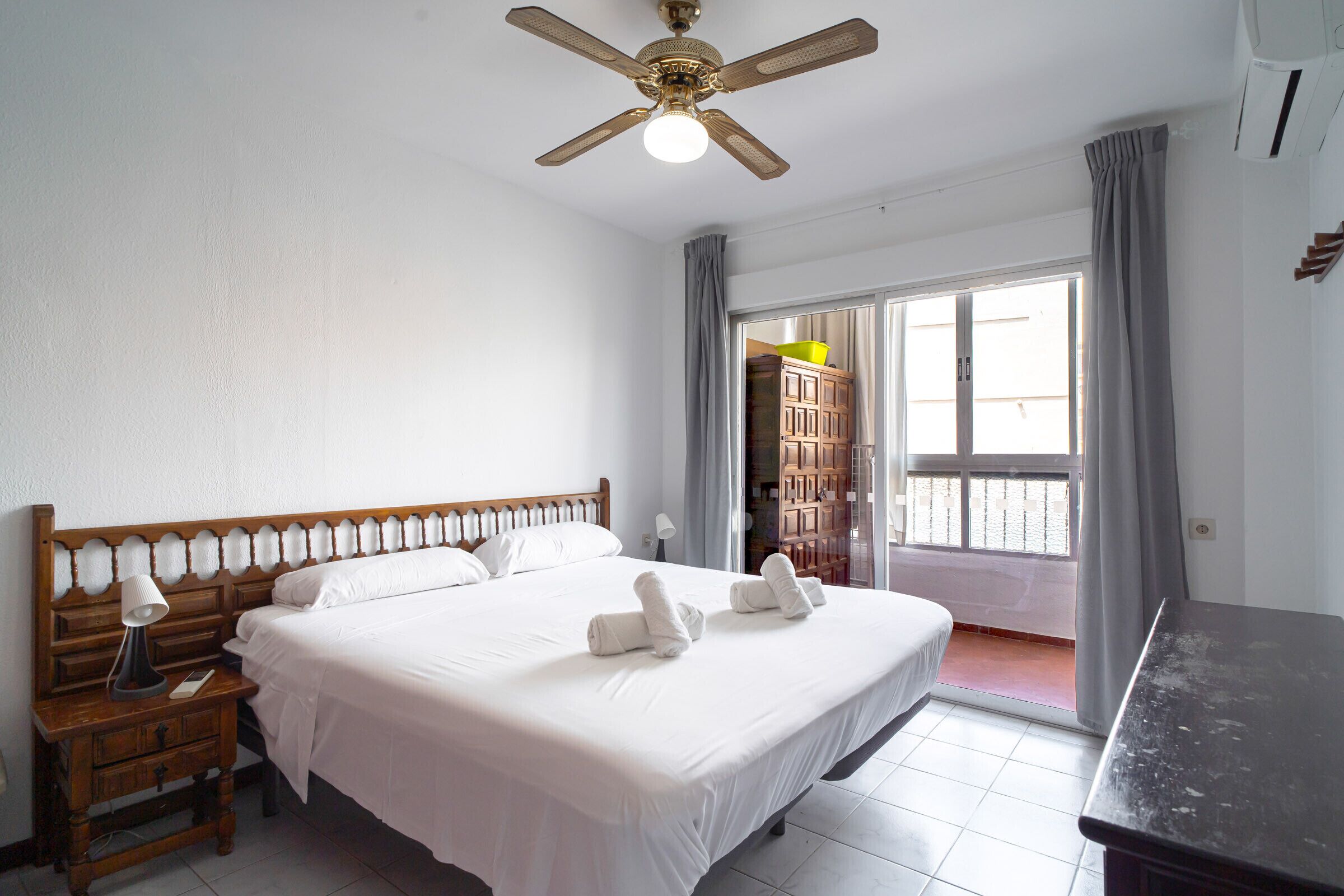 A hotel room with a large bed, a wooden nightstand, a desk, a ceiling fan, and a view of the outside through a sliding door.