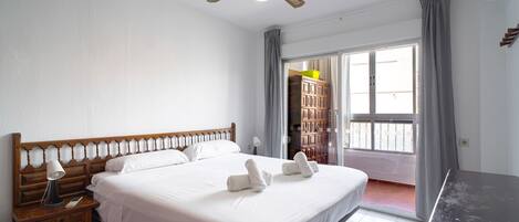 A hotel room with a large bed, a wooden nightstand, a desk, a ceiling fan, and a view of the outside through a sliding door.