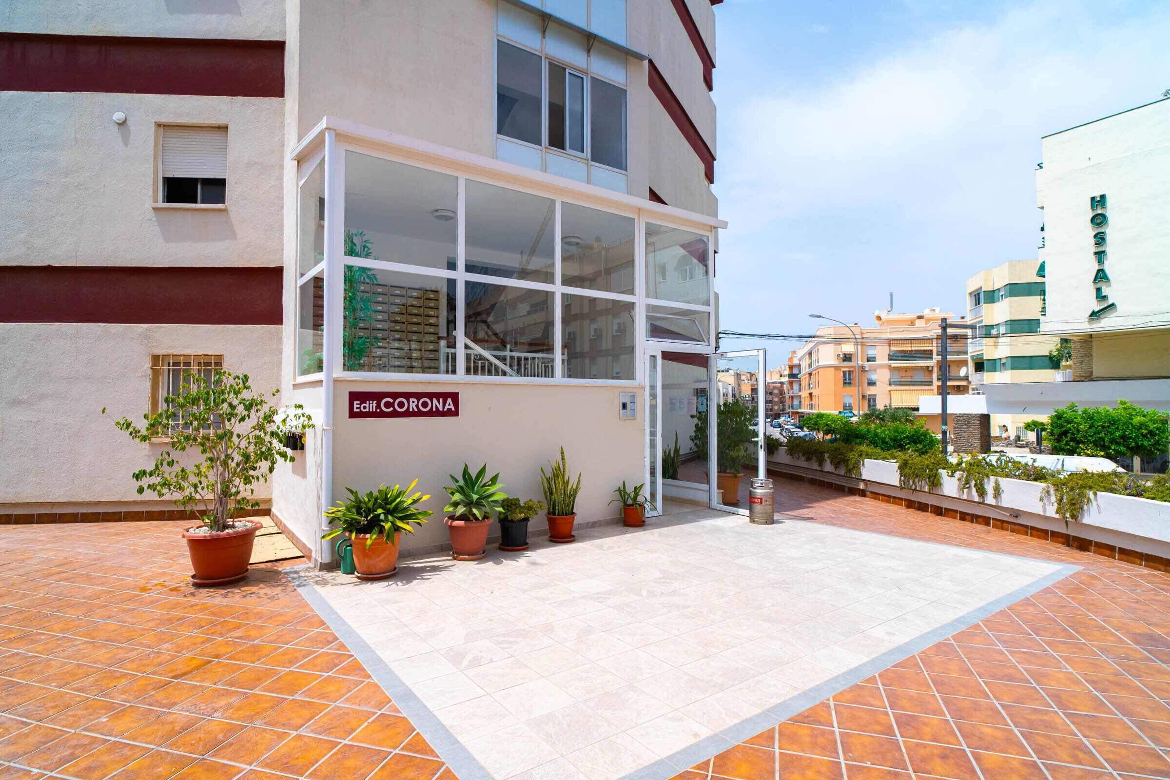 Entrada d'hotel amb un rètol que diu 'Edif. CORONA', façana de vidre i plantes en testos al terra.
