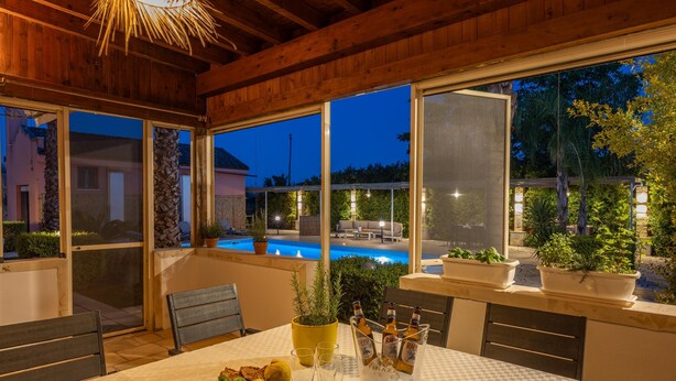 Un espacio comedor al aire libre techado con vistas a una piscina y un atardecer.