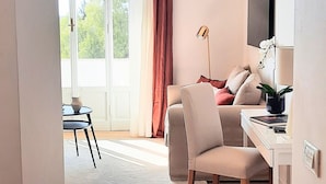 A modern living room with a sofa, a desk, and a television. There is a large window with curtains, and a view of greenery outside.