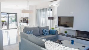 Un salon moderne avec un canapé gris, un téléviseur à écran plat et un coin repas donnant sur l’extérieur grâce à une porte vitrée.