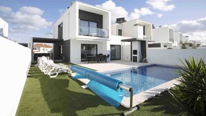 Une maison moderne à deux étages, dotée d’une piscine, d’un patio couvert et d’une pelouse aménagée avec des chaises longues.