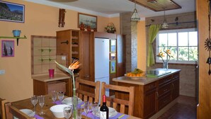 A kitchen with wooden cabinets, a refrigerator, a dining table with chairs, and a window with curtains.