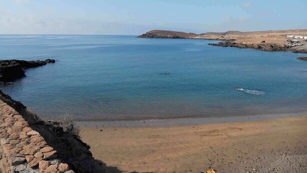 A coastal landscape with a clear sky, calm sea, rocky shoreline, and a sandy beach.
