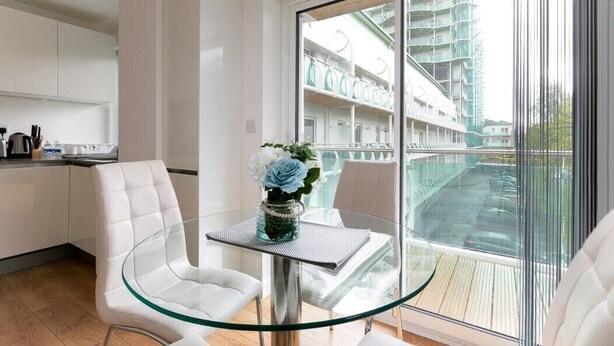 A modern room with a glass table and chairs, a view of a building with balconies, and a kitchen area with white cabinets. - Skyvillion - 1Bed Enfield Flat w/Balcony+Parking (Greater London)
