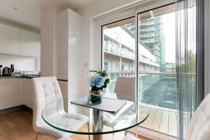A modern room with a glass table and chairs, a view of a building with balconies, and a kitchen area with white cabinets. - Skyvillion - 1Bed Enfield Flat w/Balcony+Parking (Greater London)
