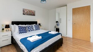 A neatly arranged bedroom with a bed, bedside tables, a nightstand, a wall-mounted lamp, and a door. - Skyvillion - 1Bed Enfield Flat w/Balcony+Parking (Greater London)