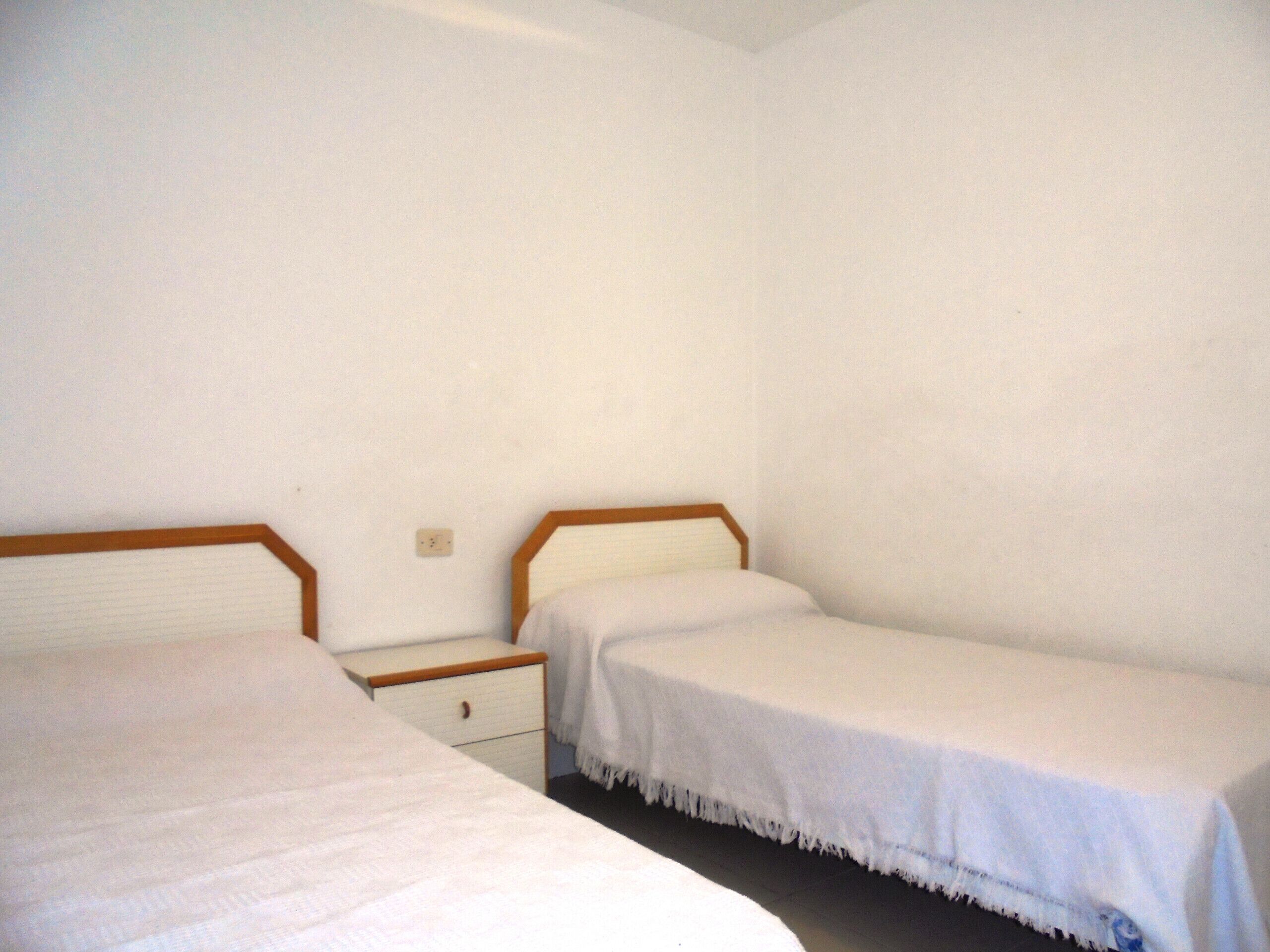 Una habitación sencilla con dos camas individuales, una mesita de noche y una pared blanca lisa.