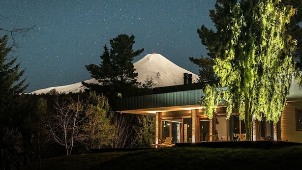 Une maison moderne dotée d’une grande façade en verre, avec une montagne en arrière-plan et un arbre au premier plan.