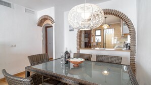 Un comedor con una mesa de vidrio rodeada de sillas de mimbre, una lámpara decorativa y una cocina visible a través de un arco.