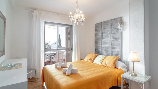 Habitación de hotel con cama, mesita de noche con lámpara, ventana con cortinas, una silla y vistas al exterior.