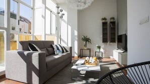 Een moderne woonkamer met een grijze bank, een salontafel en een televisie. Er zijn grote ramen waardoor natuurlijk licht binnenkomt en er uitzicht is op buiten.