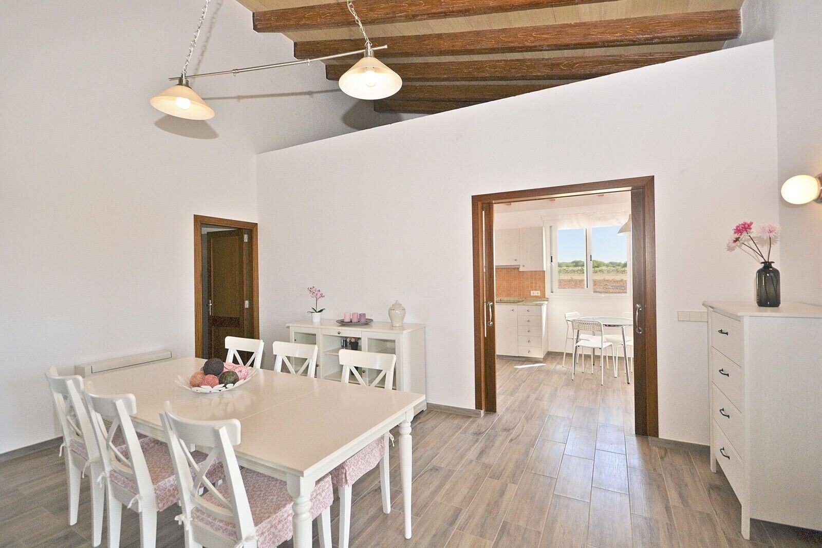 A dining area with a table and chairs, a kitchen in the background, and a wooden ceiling.