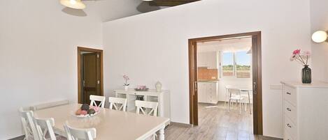 A dining area with a table and chairs, a kitchen in the background, and a wooden ceiling.