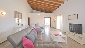 Ein großzügiges Wohnzimmer mit hoher Holzbalkendecke, einem grauen Sofa mit pinken Kissen, einem weißen Couchtisch und einem Flachbildfernseher auf einem weißen Lowboard.