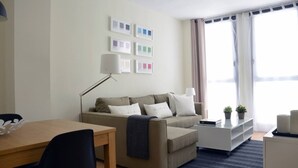A modern living room with a sofa, a dining table, and a lamp. There are framed pictures on the wall and a window with curtains.