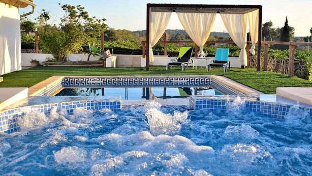 A hot tub with a tiled border, surrounded by a patio area with outdoor furniture and a pergola with draped curtains. - Can Butxaquí 160 fantastic villa with private pool and jacuzzi, air conditioning, barbecue and WiFi (Muro)