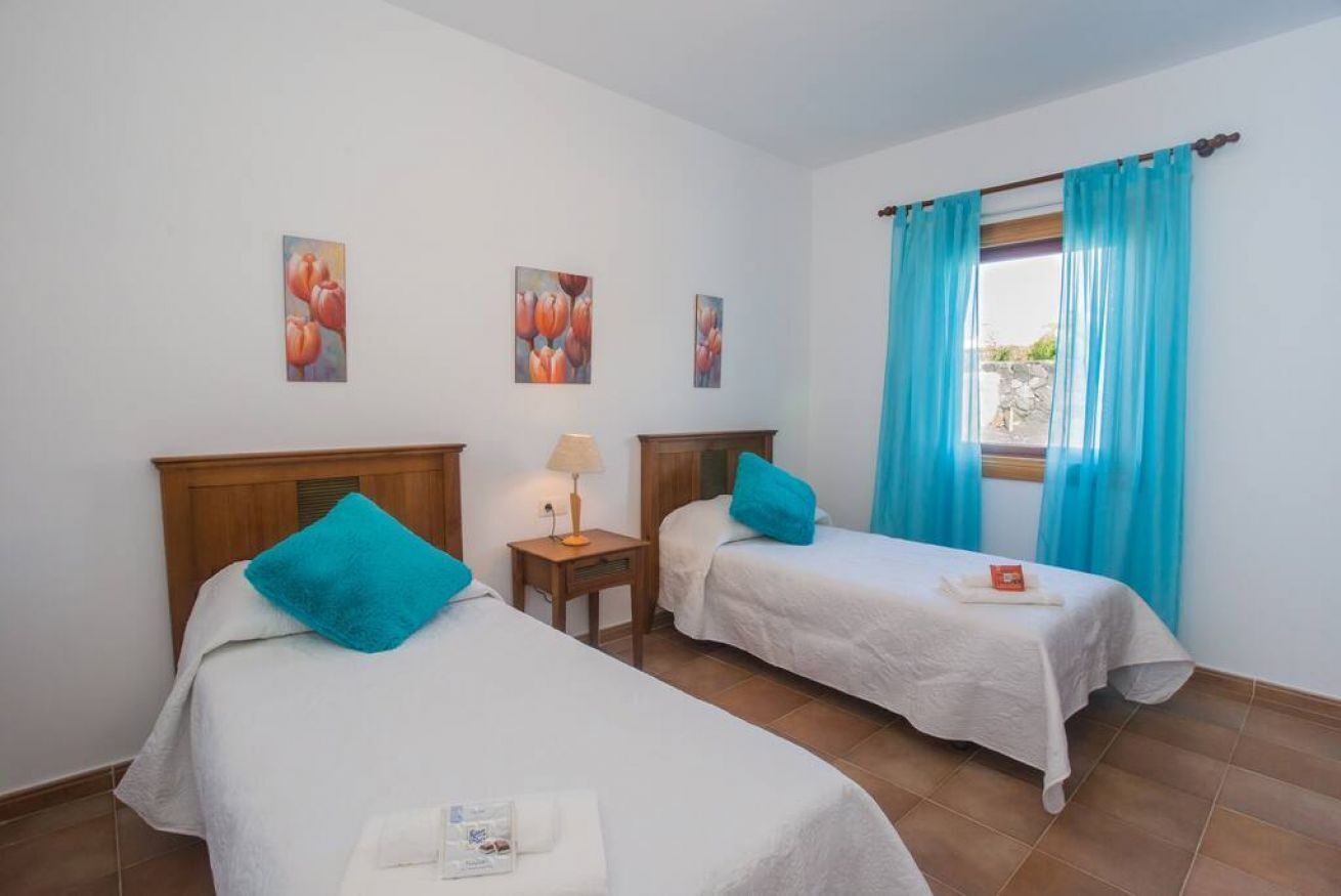 Una habitación con dos camas, cada una con cubrecama blanco y almohada azul. Hay dos cuadros en la pared, una lámpara en una mesita de noche y una ventana con cortinas azules.