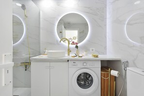 Ein modernes Badezimmer mit integrierter Waschmaschine, einem Waschbecken mit Goldarmatur und einem großen Spiegel mit runder Beleuchtung.