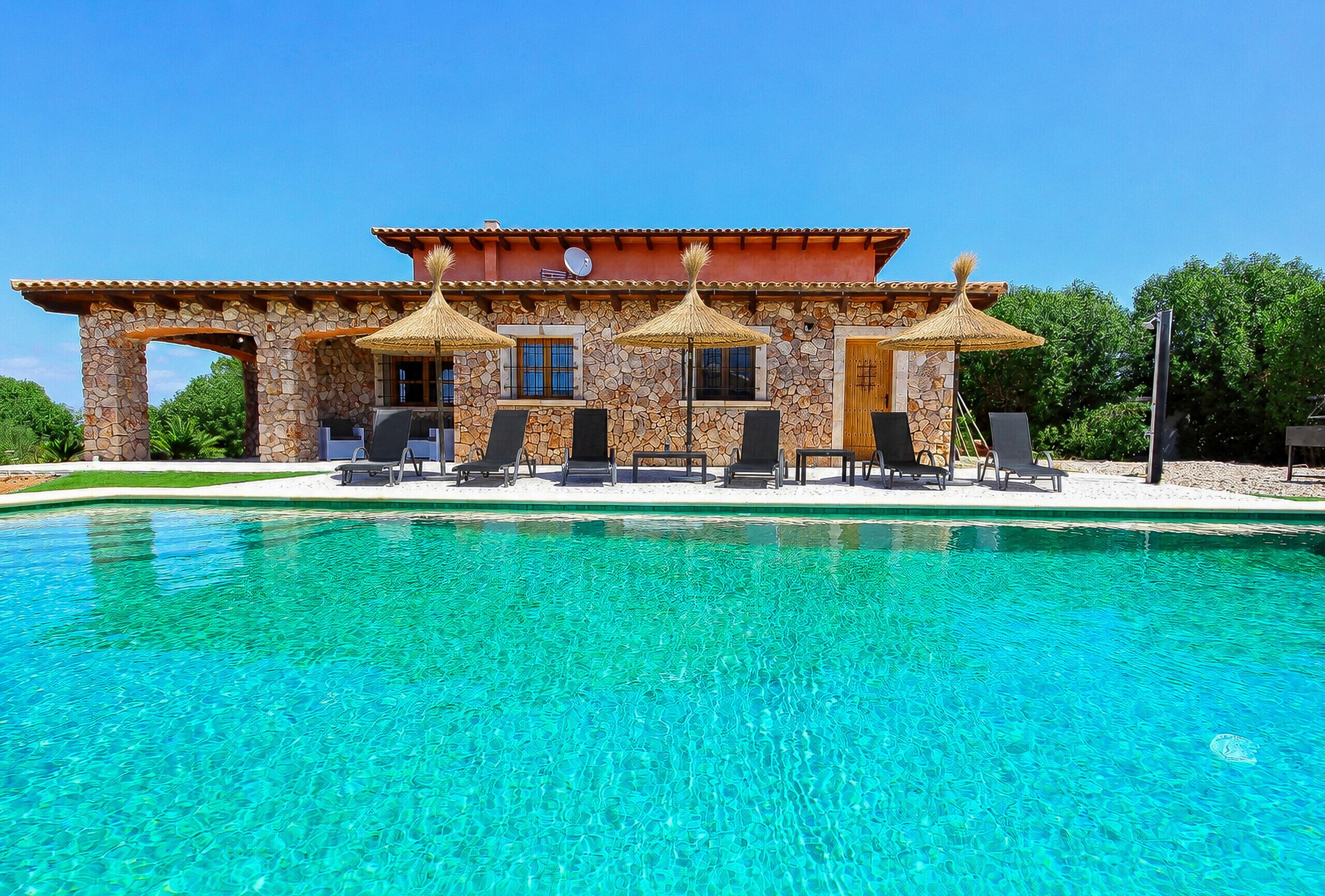 Un espace aménagé au bord de la piscine, avec un bâtiment en pierre, des sièges extérieurs et une piscine à l’eau d’un bleu limpide.