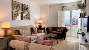 A living room with a sofa, armchairs, a coffee table, and a lamp. There are framed pictures on the wall and a view of the outside through a door.