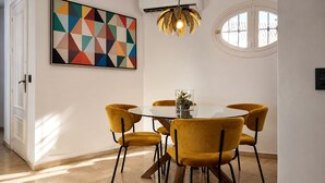 A dining area with a round table, four chairs, a geometric painting, a pendant light, and a window.