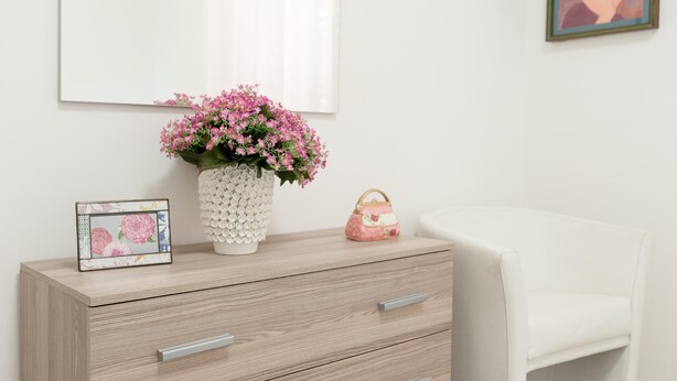 A wooden dresser with drawers, a white vase with pink flowers, a framed picture, and a pink bag.