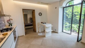 A modern kitchen with a white countertop, a sink, and a stove. There is a white bar stool and a small white table with a vase of flowers. A large window provides a view of greenery outside. - Apartment on the Amalfi Coast PARKING SPACE AND POOL (Salerno)