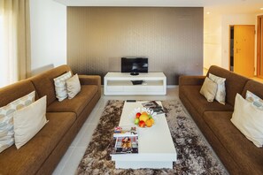 A living room with two brown sofas, a white coffee table with magazines and fruit, and a TV on a white stand.