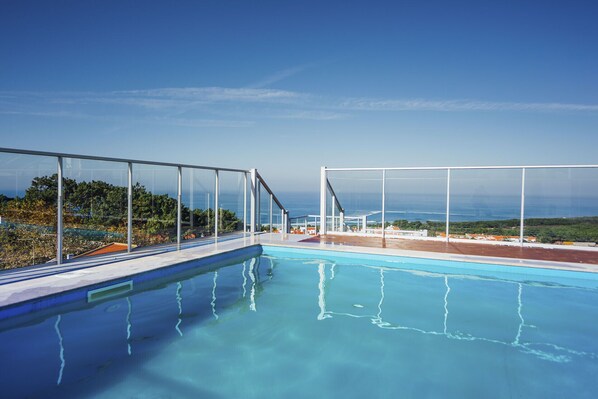 A clear blue swimming pool with a glass barrier, a view of the ocean, and a clear sky.