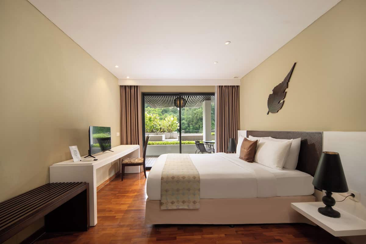 A modern hotel room with a large bed, a desk with a flat-screen TV, a bench, and a view of greenery outside.