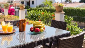 A table with fruit and drinks, wicker chairs, and a view of a house and garden.