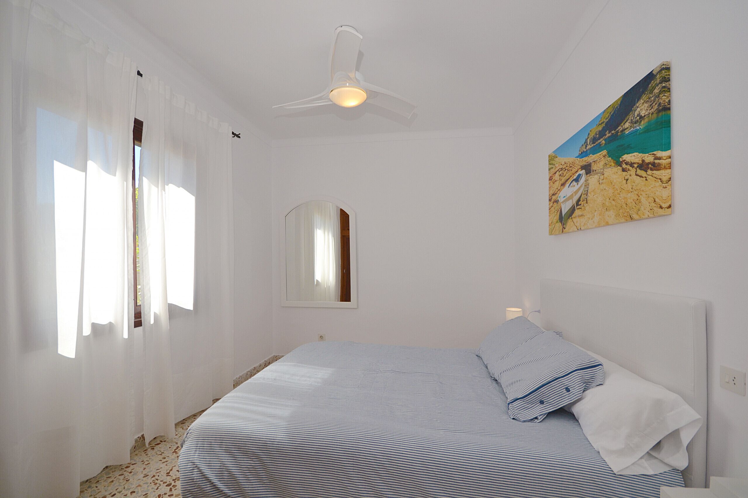 A bedroom with a bed, a ceiling fan, a window with sheer curtains, a wall-mounted mirror, and a painting of a coastal scene.