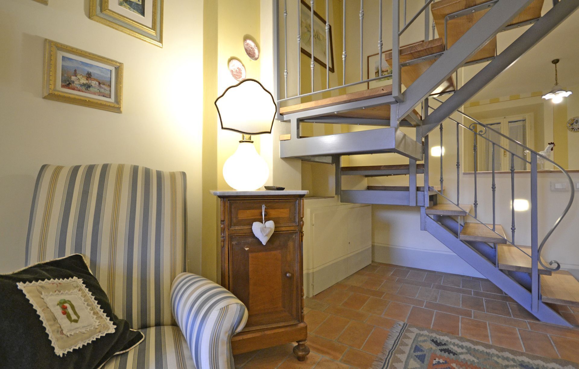 Una sala de estar con un sillón a rayas, un mueble de madera con un adorno con forma de corazón, una escalera con barandillas de metal y un suelo de baldosas.