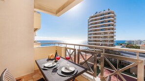 Un balcón con una mesa lista para dos personas, con vistas al mar y un edificio alto al fondo.