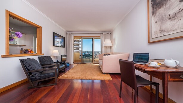 Une chambre d’hôtel avec un plancher en bois, un canapé, un bureau avec un ordinateur portable, une chaise, une table basse, un miroir, une fenêtre donnant sur un balcon et un tableau encadré au mur.