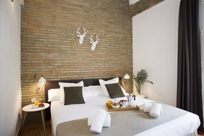 A hotel room with a brick wall, a bed with white bedding, two bedside tables with lamps, a tray with breakfast items, and a plant.