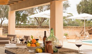 An outdoor dining area with a table set for two, featuring a bottle, glasses, and a bowl of fruit.