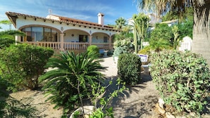 Una casa de dos pisos amb un balcó, envoltada de vegetació abundant i cactus.