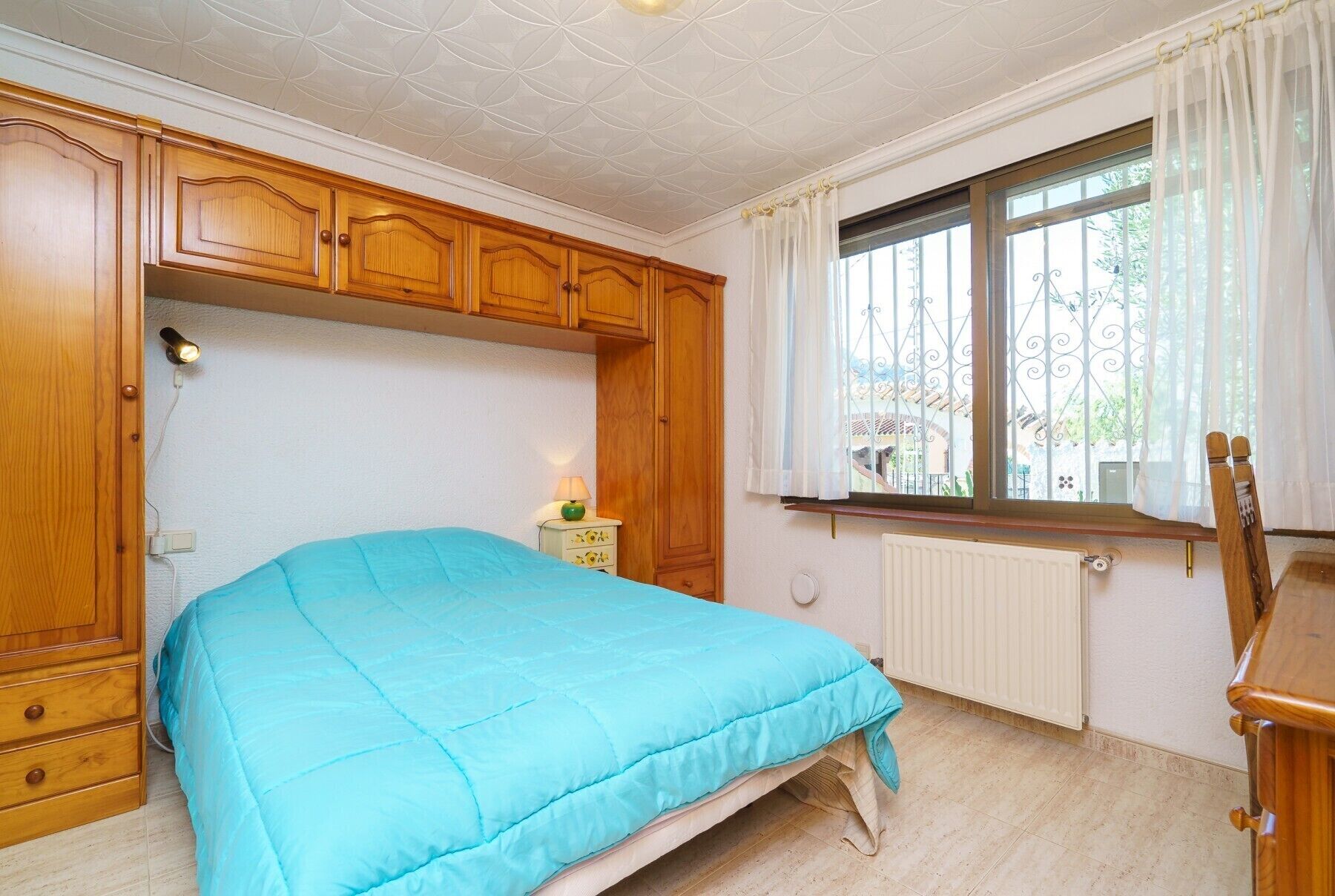 Une chambre à coucher avec un lit, une armoire en bois, une fenêtre avec des rideaux, un radiateur et une petite table avec une lampe.
