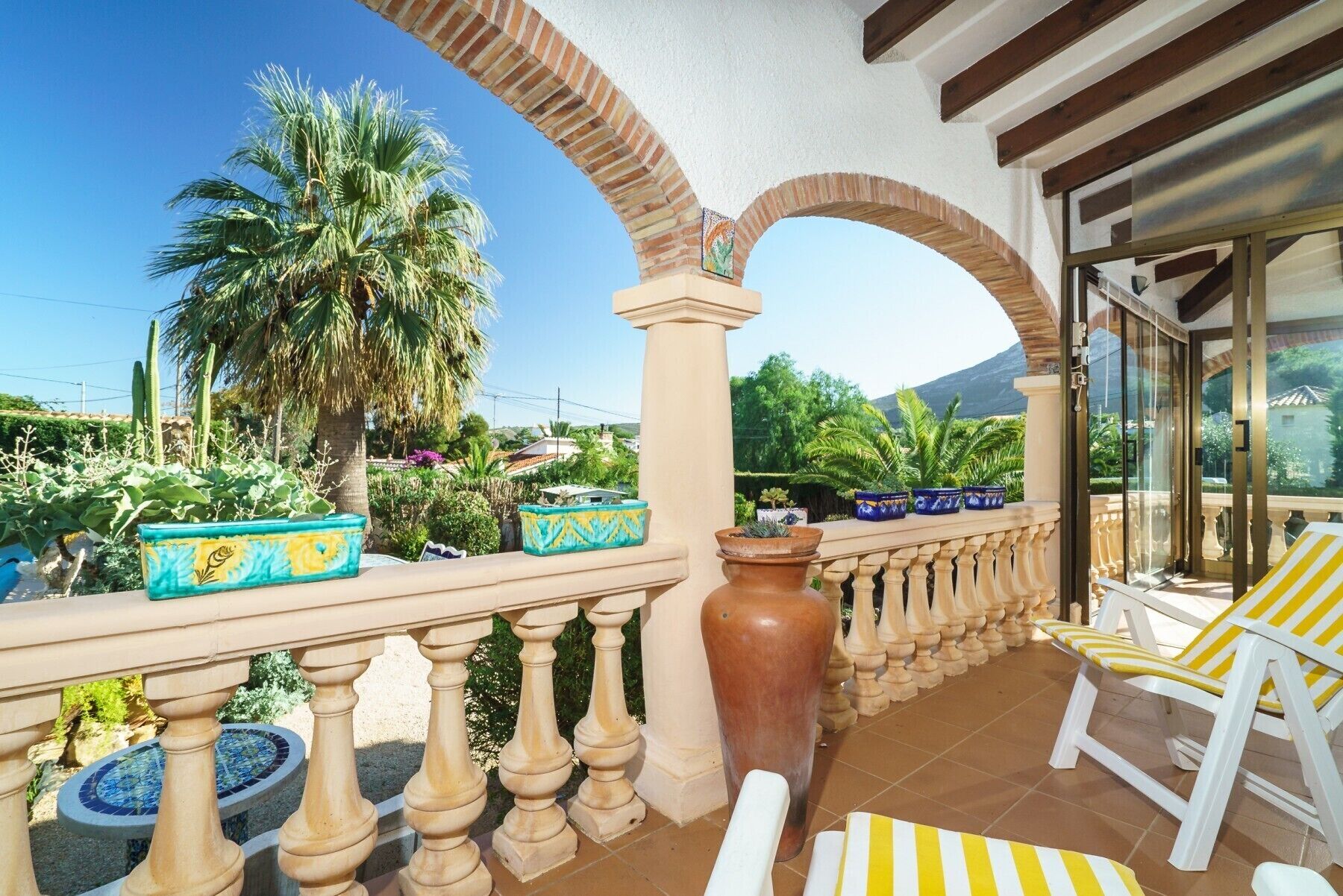 A balcony with a view of palm trees and a mountain, featuring a tiled floor, a large vase, and striped chairs.