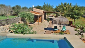 Ein Schwimmbecken mit Holzterrasse und Liegestühlen, umgeben von einer rustikalen Außenanlage mit einem Reetdach und viel Grün.