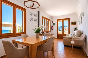 Un comedor con mesa y sillas de madera, una planta en maceta y vistas al mar a través de amplios ventanales.