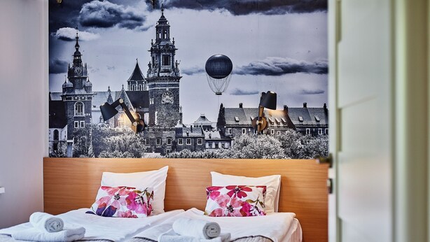 A hotel room with a bed featuring two pillows, a wooden headboard, and a wall mural of a cityscape with a hot air balloon. - Irena and Anatol Apartment - 5 min walk to Krakow’s Main Square! Sleeps 4!  (Kraków)