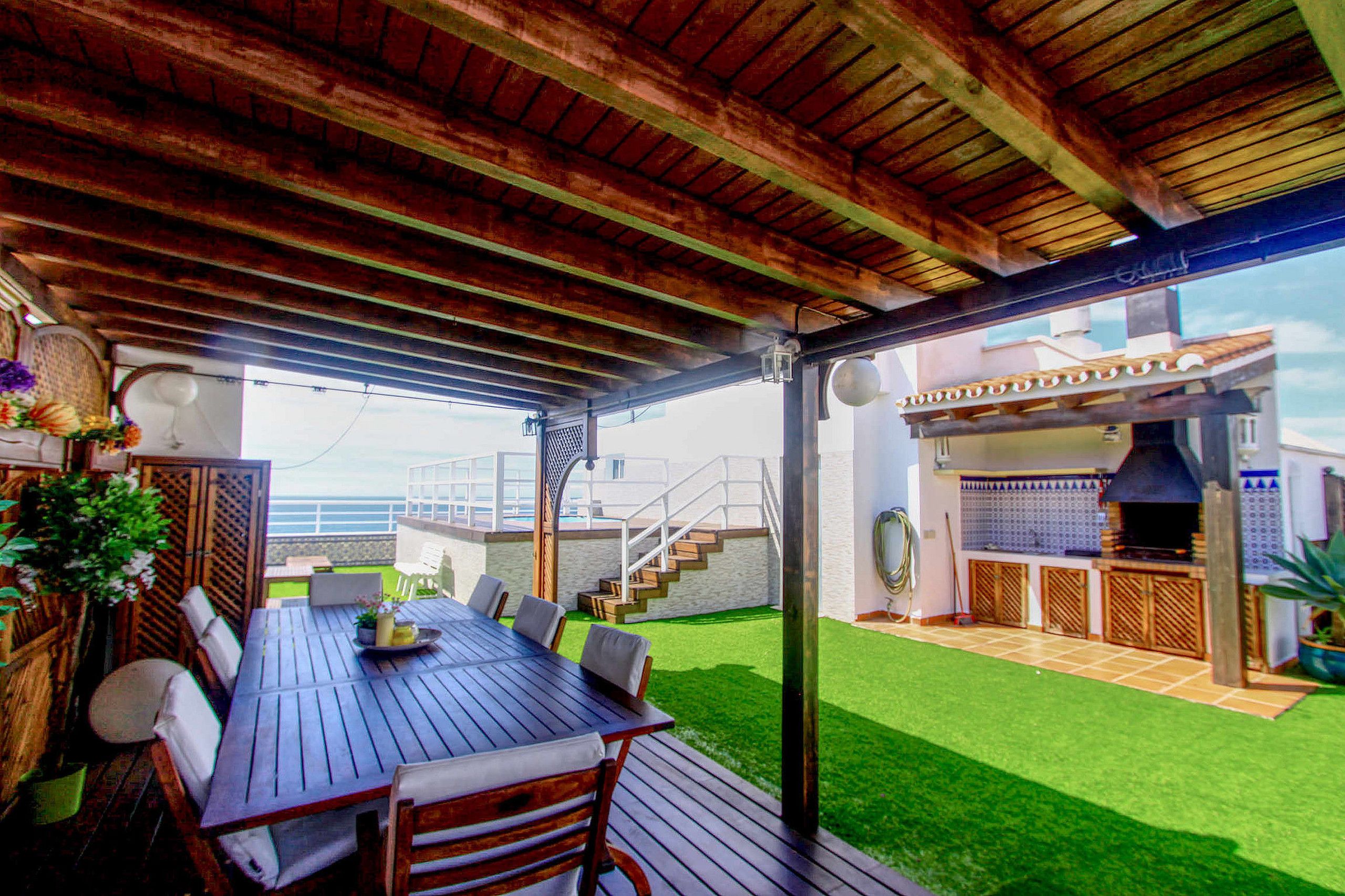 An outdoor patio with a wooden table and chairs, a barbecue area, and a view of the ocean.